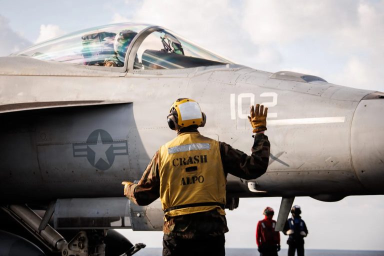 A deck crew member in a yellow safety vest signaling to a fighter jet pilot on an aircraft carrier.