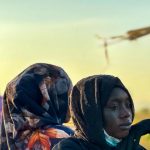 A close-up portrait of a Sudanese woman wearing a black hijab with a somber expression.