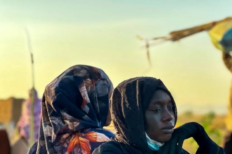 A close-up portrait of a Sudanese woman wearing a black hijab with a somber expression.