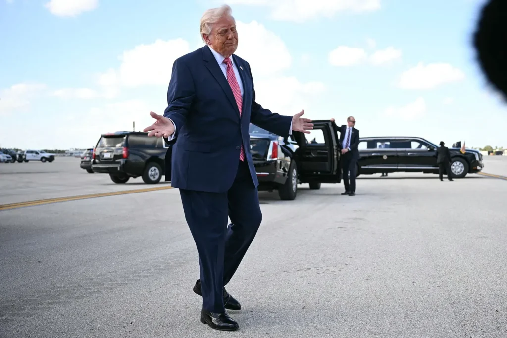Donald Trump gesturing with open arms while walking on an airport tarmac near official vehicles.