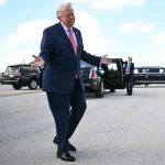 Donald Trump gesturing with open arms while walking on an airport tarmac near official vehicles.