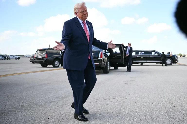 Donald Trump gesturing with open arms while walking on an airport tarmac near official vehicles.