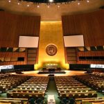 Wide-angle view of the empty United Nations General Assembly hall in New York with the UN emblem on the golden front wall.