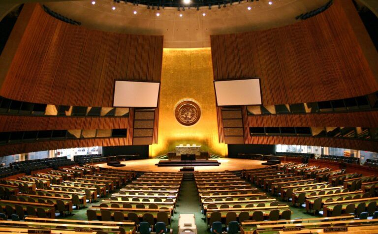 Wide-angle view of the empty United Nations General Assembly hall in New York with the UN emblem on the golden front wall.