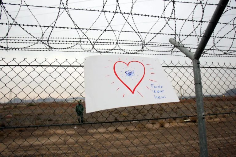 A hand-drawn sign on a chain-link fence with a heart and an atom symbol, reading "Fordo is our heart," near a military facility.