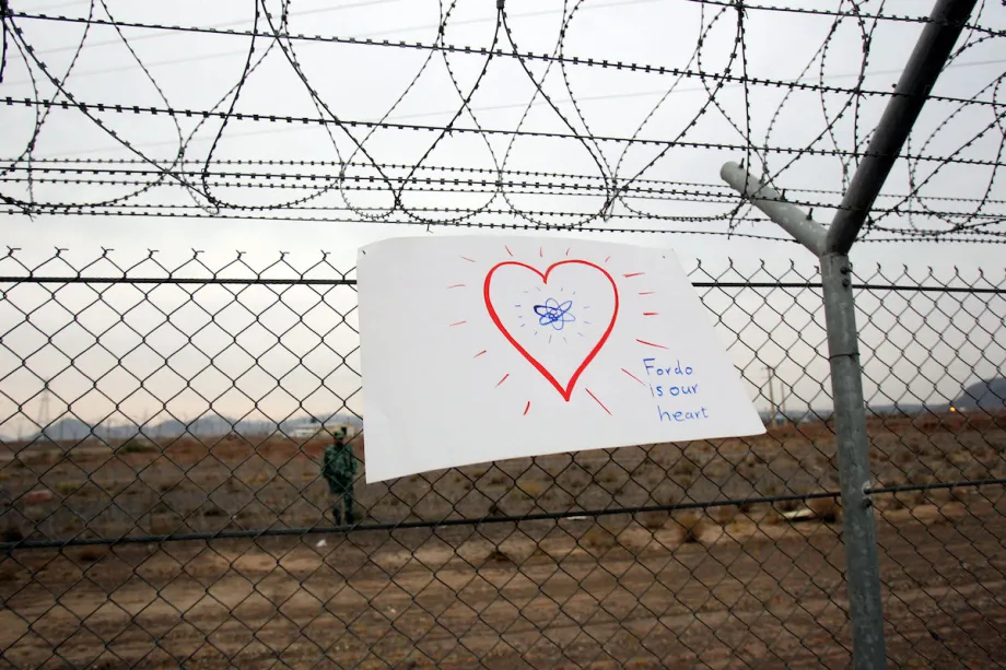 A hand-drawn sign on a chain-link fence with a heart and an atom symbol, reading "Fordo is our heart," near a military facility.