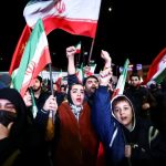 A crowd of people, including women and children, waving Iranian flags during a nighttime demonstration.