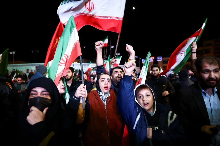 A crowd of people, including women and children, waving Iranian flags during a nighttime demonstration.