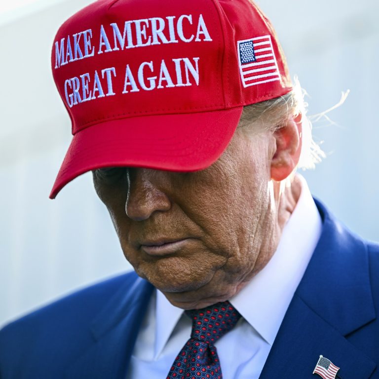 Close-up portrait of Donald Trump wearing a red "Make America Great Again" hat looking downwards.