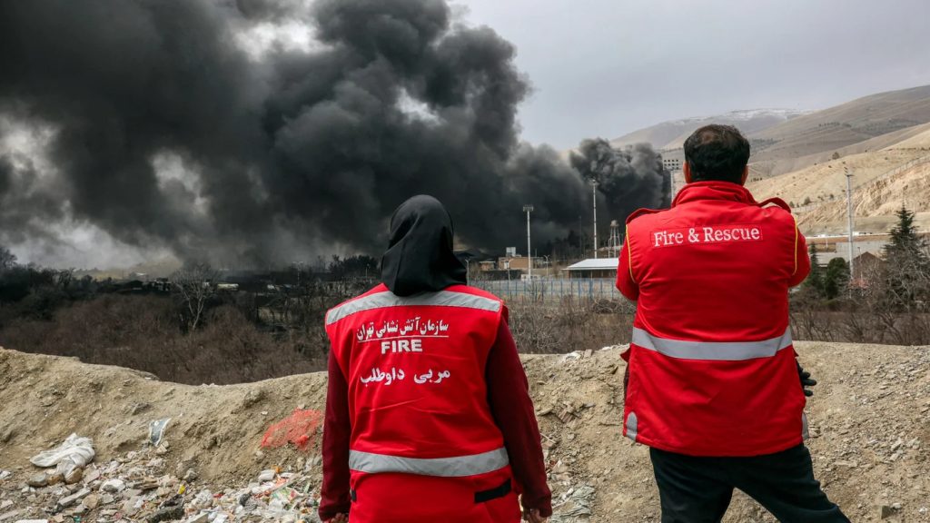Two Iranian firefighters in red vests looking at thick black smoke rising from a distant facility.