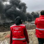 Two Iranian firefighters in red vests looking at thick black smoke rising from a distant facility.