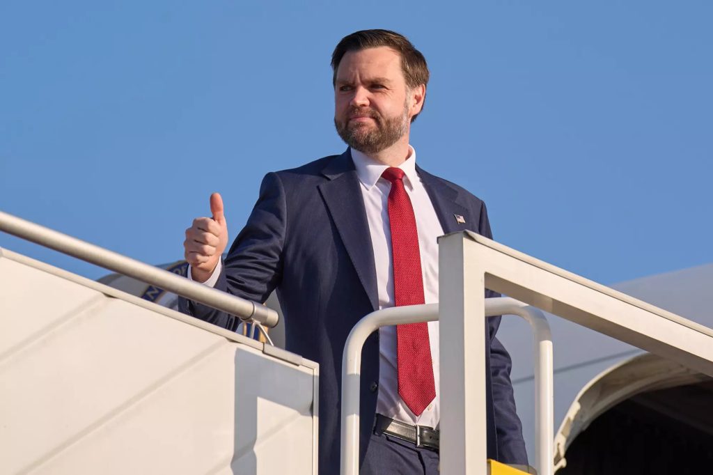 JD Vance giving a thumbs-up while standing on the stairs of an airplane against a clear blue sky.