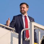 JD Vance giving a thumbs-up while standing on the stairs of an airplane against a clear blue sky.