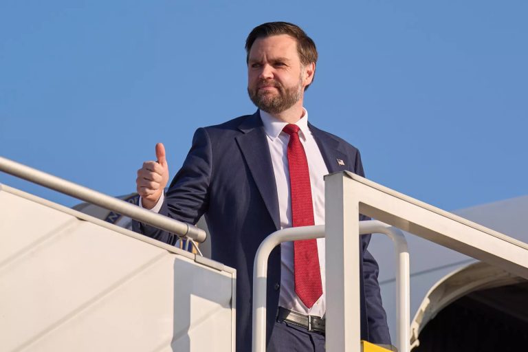JD Vance giving a thumbs-up while standing on the stairs of an airplane against a clear blue sky.