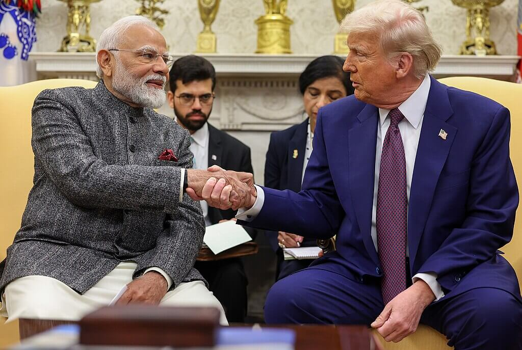 Indian Prime Minister Narendra Modi and President Donald Trump shaking hands while seated in the Oval Office, with aides and a fireplace in the background.