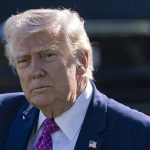 A portrait of Donald Trump looking directly at the camera with a serious expression, wearing a blue suit and purple tie.