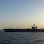A silhouette of a US Navy aircraft carrier at sea during sunset with a fighter jet taking off.