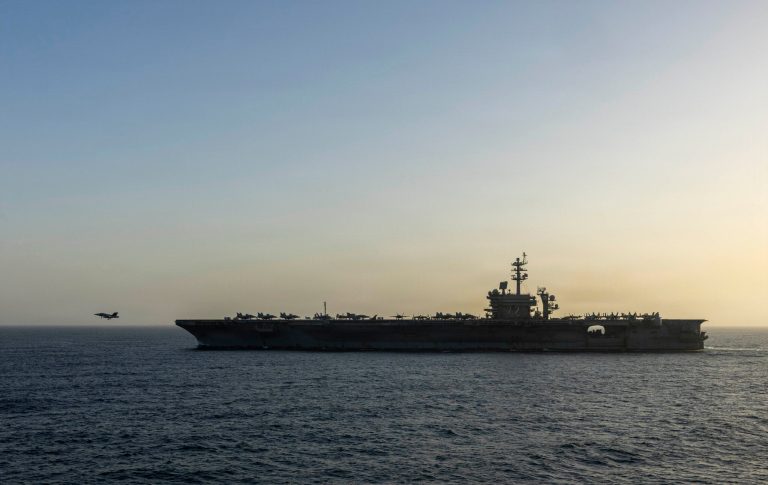 A silhouette of a US Navy aircraft carrier at sea during sunset with a fighter jet taking off.