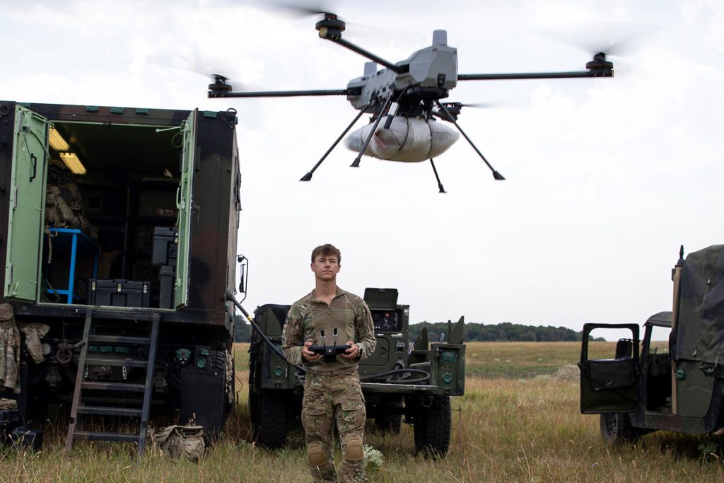 A military operator in camouflage uniform standing in a field, controlling a large grey quadcopter drone that is hovering in the air near a tactical mobile command vehicle.