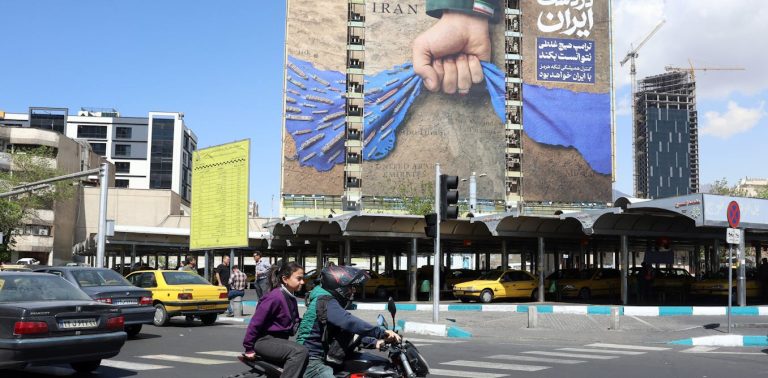 A street scene in Tehran featuring a large political billboard showing a hand controlling the Strait of Hormuz.