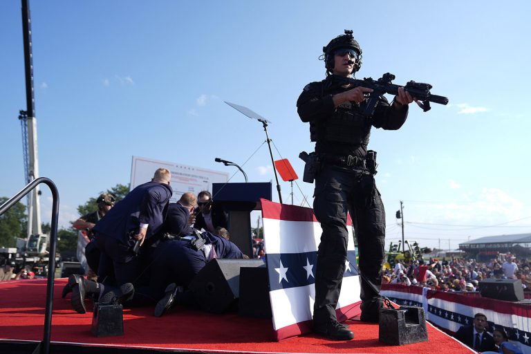 Secret Service agents and tactical law enforcement officers securing a stage during an emergency incident at a political rally, with an officer in tactical gear holding a rifle in the foreground.