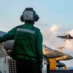 A flight deck crew member in a green vest watching a fighter jet take off or land on an aircraft carrier.