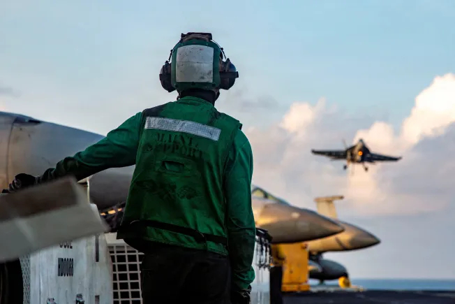 A flight deck crew member in a green vest watching a fighter jet take off or land on an aircraft carrier.