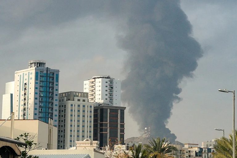 Large plumes of dark gray smoke rising behind urban apartment buildings under an overcast sky.