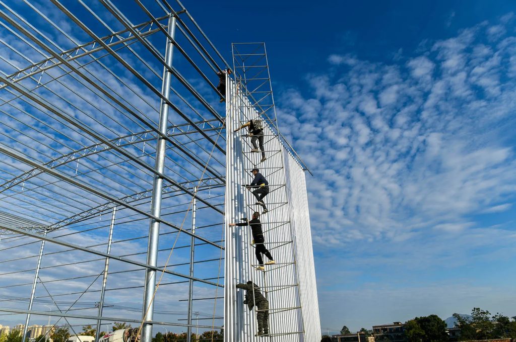 Workers climbing a high metal scaffolding to install white panels on a large building frame.