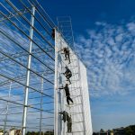 Workers climbing a high metal scaffolding to install white panels on a large building frame.