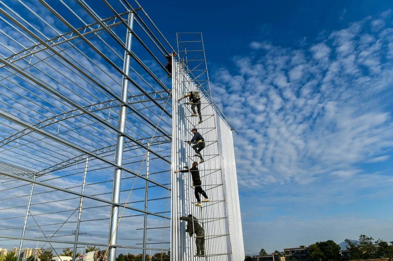 Workers climbing a high metal scaffolding to install white panels on a large building frame.
