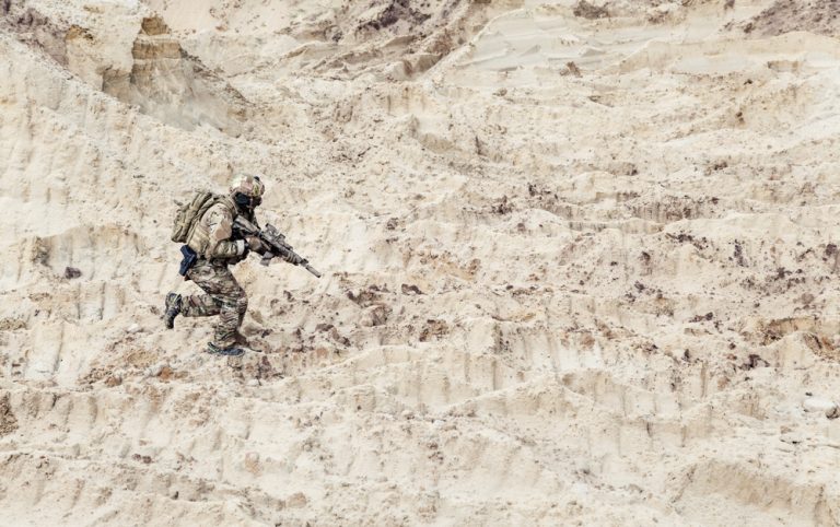 A soldier in full camouflage gear and tactical equipment running across a sandy, uneven terrain.