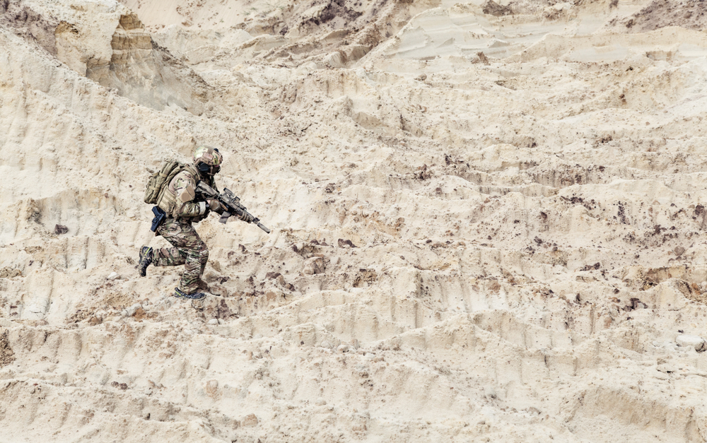 A soldier in full camouflage gear and tactical equipment running across a sandy, uneven terrain.