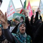 A crowd of Iranian women in black chadors protesting and holding portraits of Ayatollah Khamenei and Iranian flags.