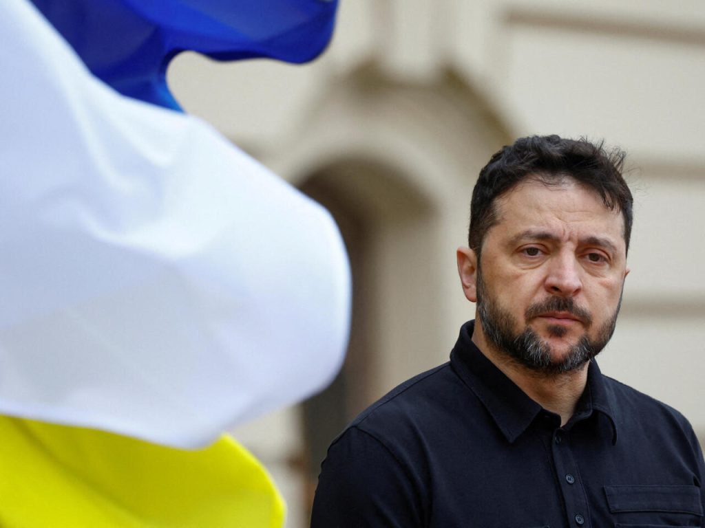 Close-up portrait of President Volodymyr Zelenskyy looking contemplative with a blurred Ukrainian flag in the foreground.