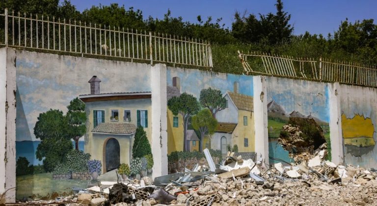 A damaged mural painted on a wall in Lebanon showing a peaceful house scene, with rubble in the foreground.