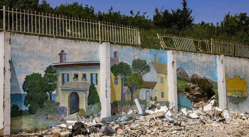 A damaged mural painted on a wall in Lebanon showing a peaceful house scene, with rubble in the foreground.