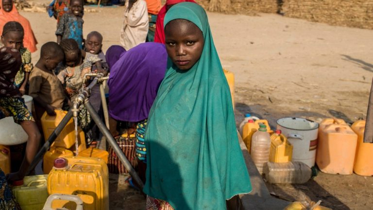 A young girl wearing a hijab standing near a water tap surrounded by yellow water containers in a humanitarian setting.