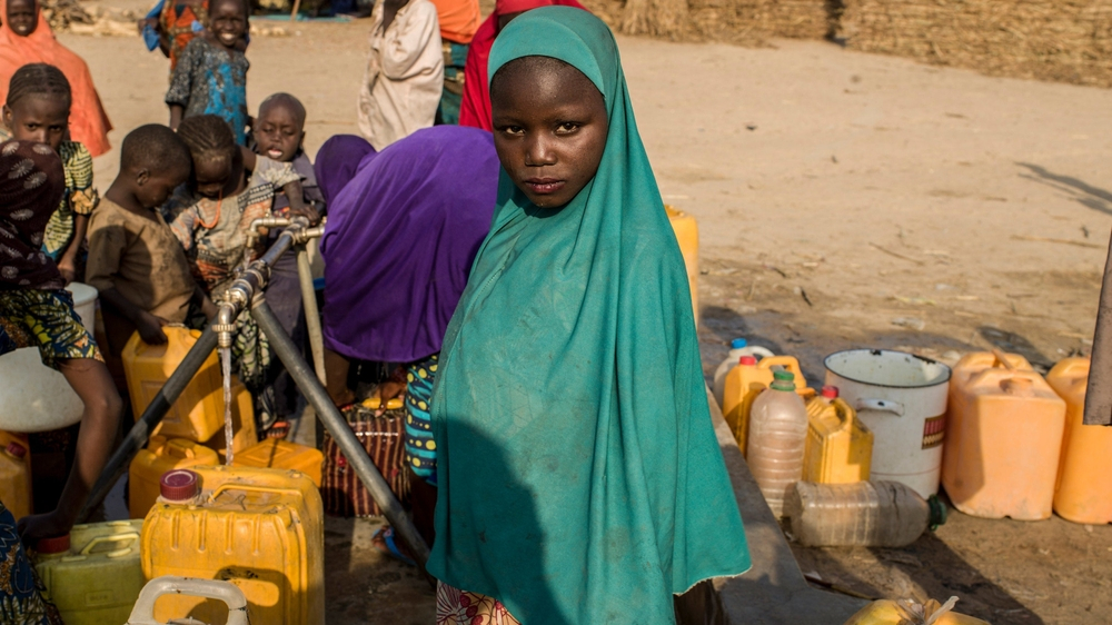 A young girl wearing a hijab standing near a water tap surrounded by yellow water containers in a humanitarian setting.