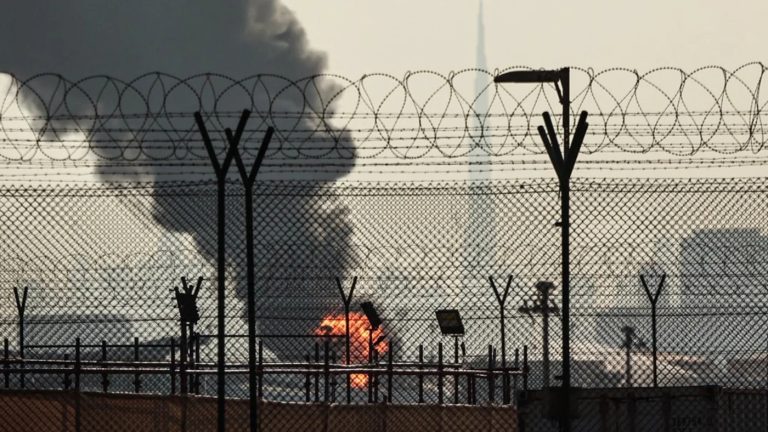A fire and thick black smoke rising from an industrial facility behind a barbed-wire fence in the Middle East.
