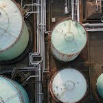 Aerial top-down view of large industrial oil storage tanks and a network of white pipelines.
