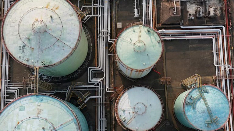 Aerial top-down view of large industrial oil storage tanks and a network of white pipelines.