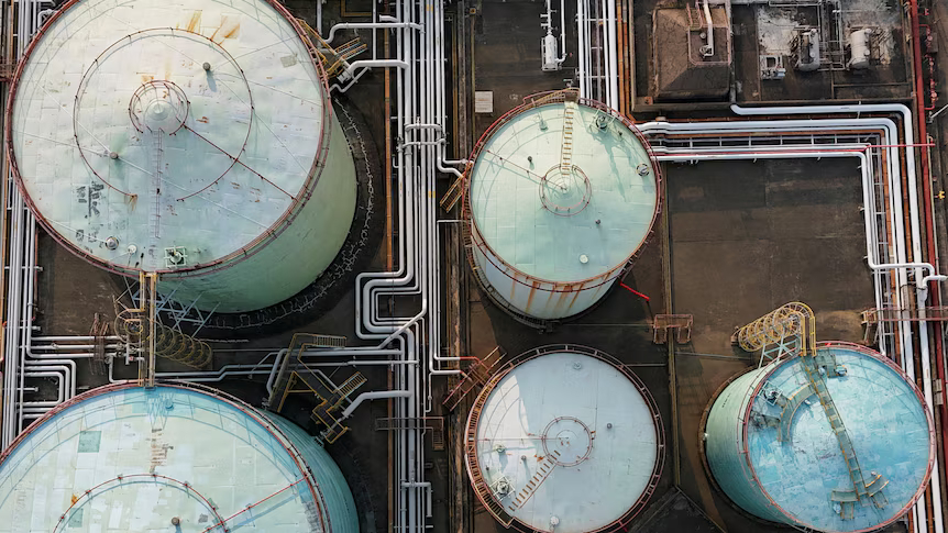Aerial top-down view of large industrial oil storage tanks and a network of white pipelines.