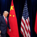 Donald Trump walking past American and Chinese flags with a somber expression.