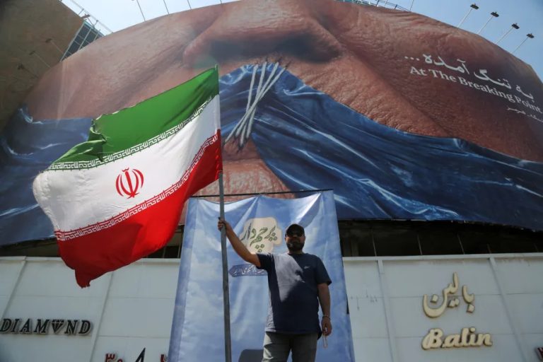 A man holding a large Iranian flag in front of a massive billboard showing a close-up of a human face with lips tied together and text that reads "At The Breaking Point".