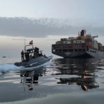 Military speedboat approaching a large container ship at dusk.