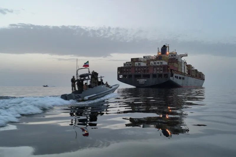 Military speedboat approaching a large container ship at dusk.