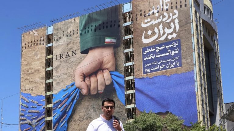 A man holding a microphone in front of a massive building mural depicting a fist gripping the Strait of Hormuz and maritime shipping lanes.
