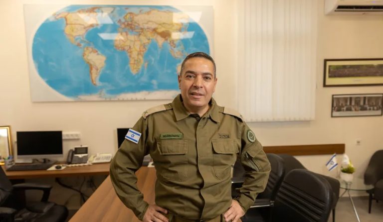 An Israeli military official in uniform standing in an office in front of a world map.