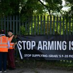 Protesters holding a large black banner that reads "STOP ARMING ISRAEL" near a metal fence with police officers nearby.